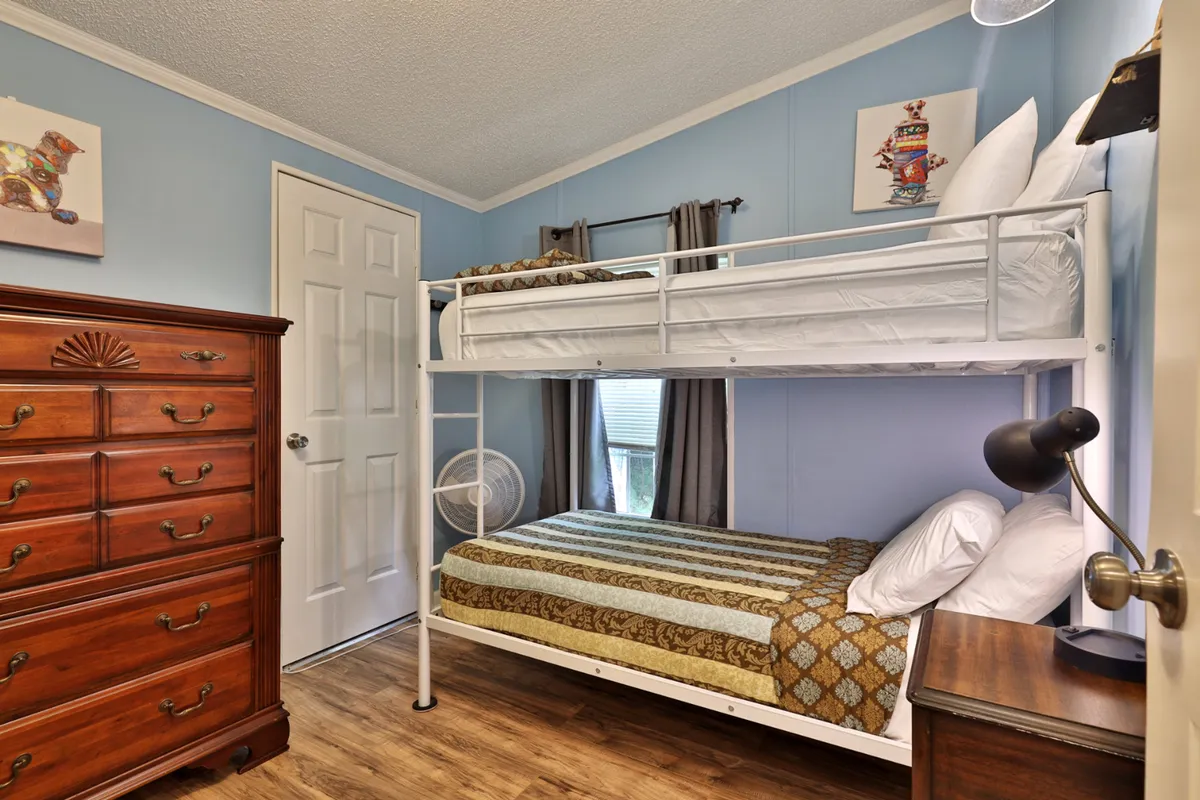 A cheerful bunk room with a sturdy bunk bed, chest of drawers, playful wall art, and a window with curtains. The inviting space is perfect for children or the young at heart, with thoughtful touches that make it feel cozy and fun.