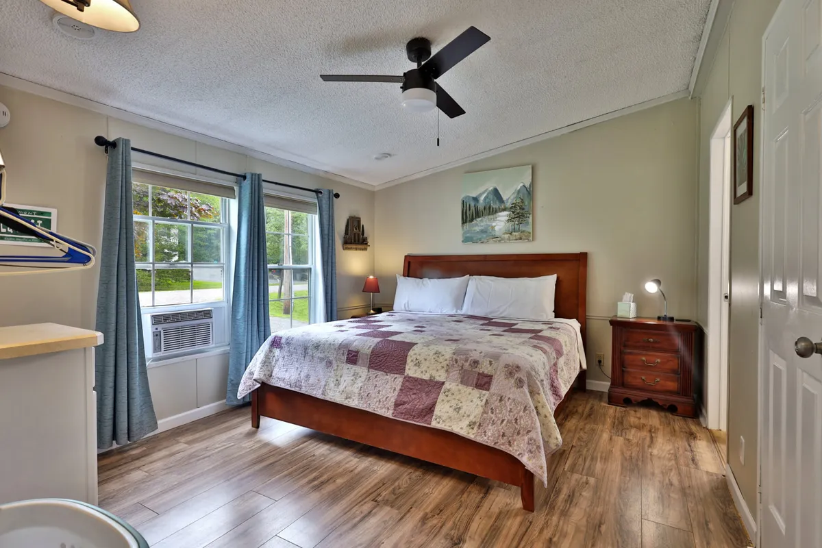 A comfortable bedroom featuring a bed with a cozy quilt, a window with curtains letting in natural light, artwork on the wall, and a nightstand with a lamp. The mood is peaceful and inviting, ideal for restful sleep.