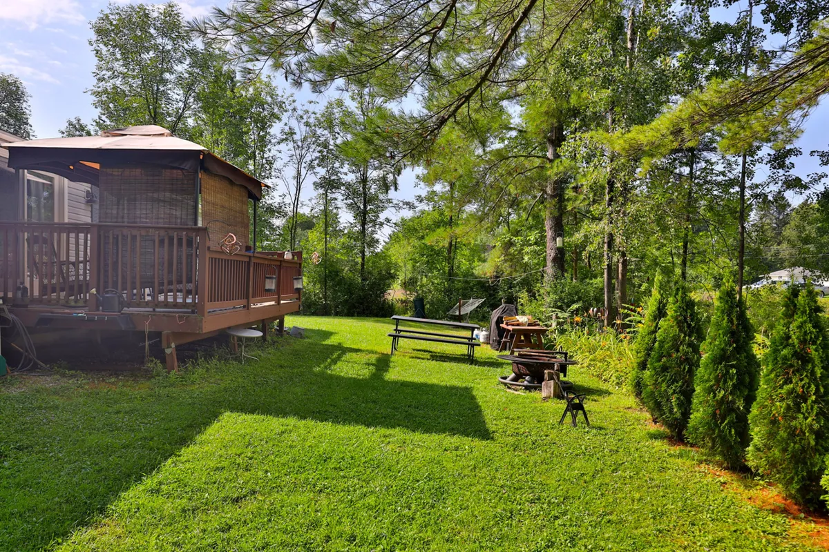 An inviting deck area overlooking a green lawn, with a large fire pit surrounded by benches and mature trees. The setup suggests a perfect spot for outdoor dining, relaxing, and gathering around the fire as day turns to night.