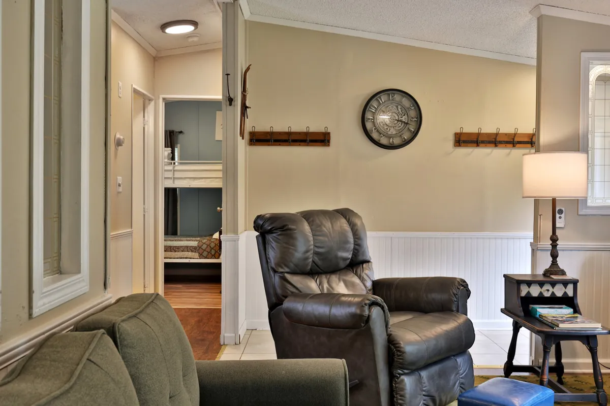 A cozy living room space with a plush recliner chair, a wall clock, a wooden side table, and a view into the bedroom through an open doorway. The atmosphere is warm and inviting, perfect for relaxing, reading, or spending time with loved ones during autumn or winter months.