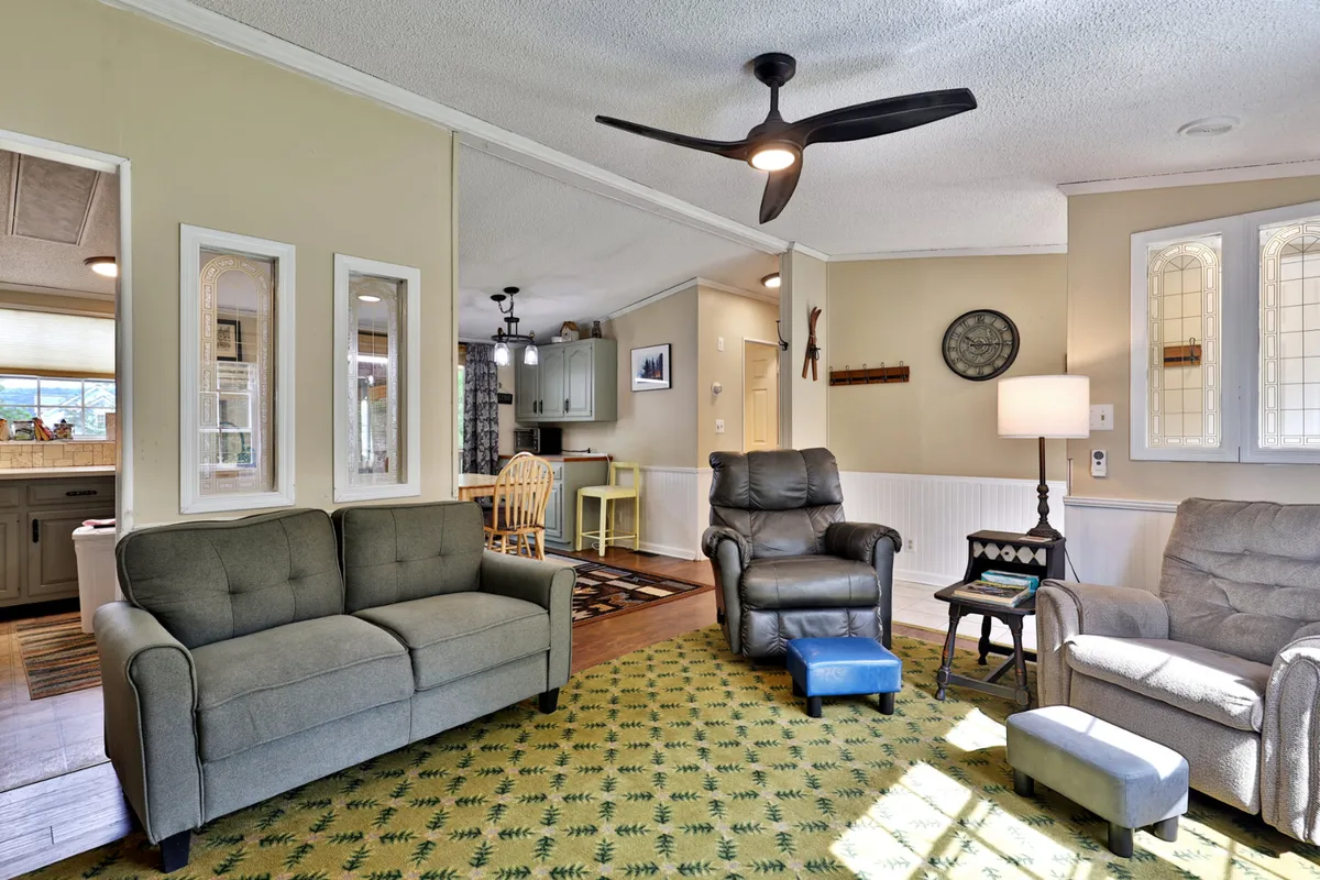 A welcoming view of the living room, featuring a comfortable seating area arranged around a television. Light filters in through nearby windows, and a textured green carpet adds warmth to the space. The dining area is visible in the background, demonstrating the open layout connecting the living and dining areas.