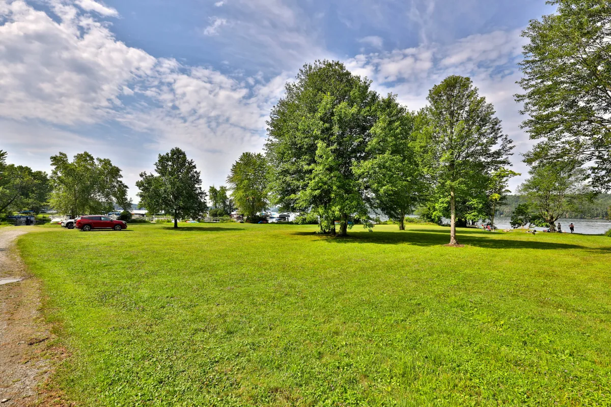 A grassy, tree-lined yard with a distant view of water, cars parked, and the suggestion of nearby recreational activity. The natural surroundings hint at space for picnicking, fishing, and unwinding in an outdoor setting.