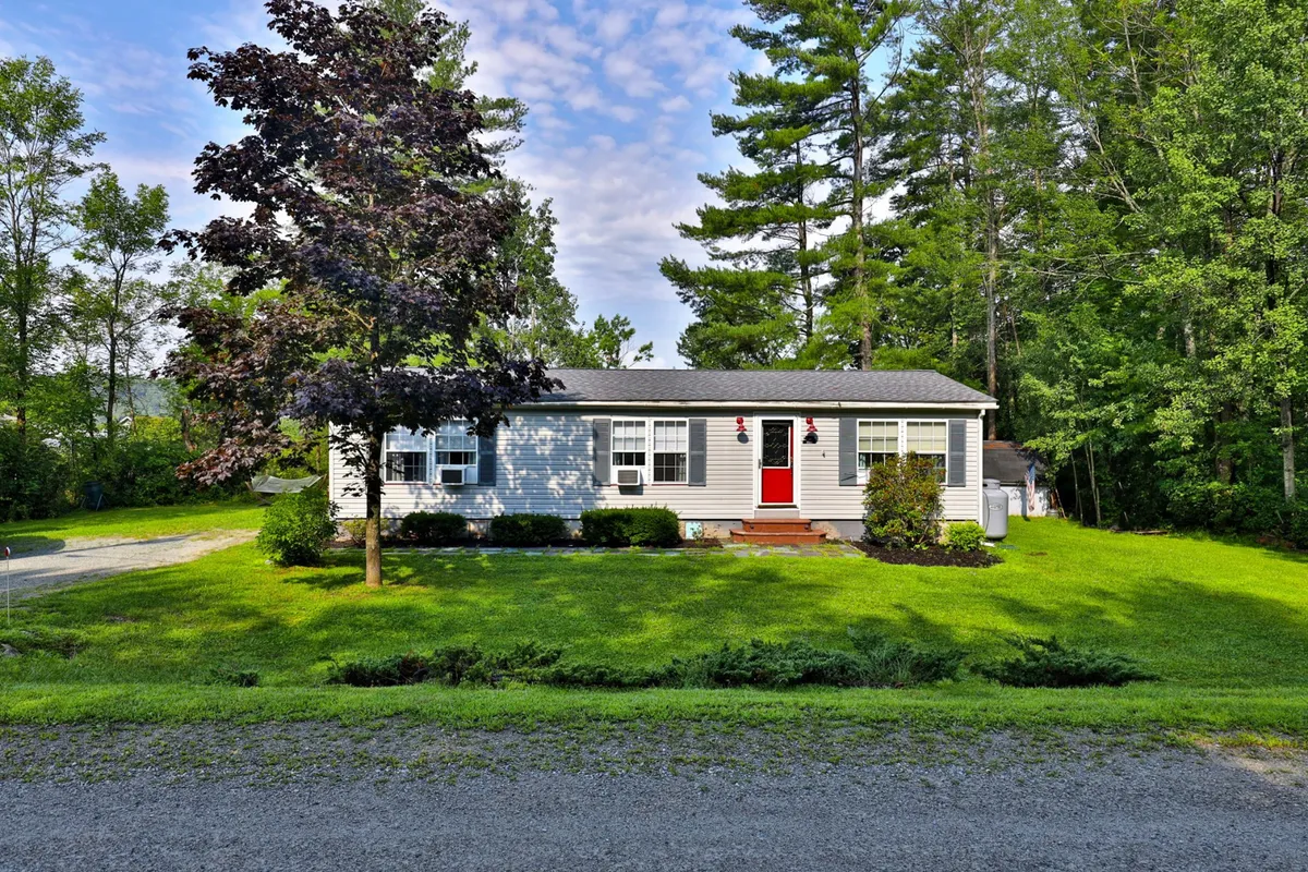 This image showcases the charming exterior of a Vermont lakeside bungalow, featuring a vibrant red front door, a lush manicured lawn perfect for outdoor activities, and mature trees offering privacy and a sense of seclusion. The inviting porch hints at leisurely afternoons spent watching sunsets or gathering with loved ones.